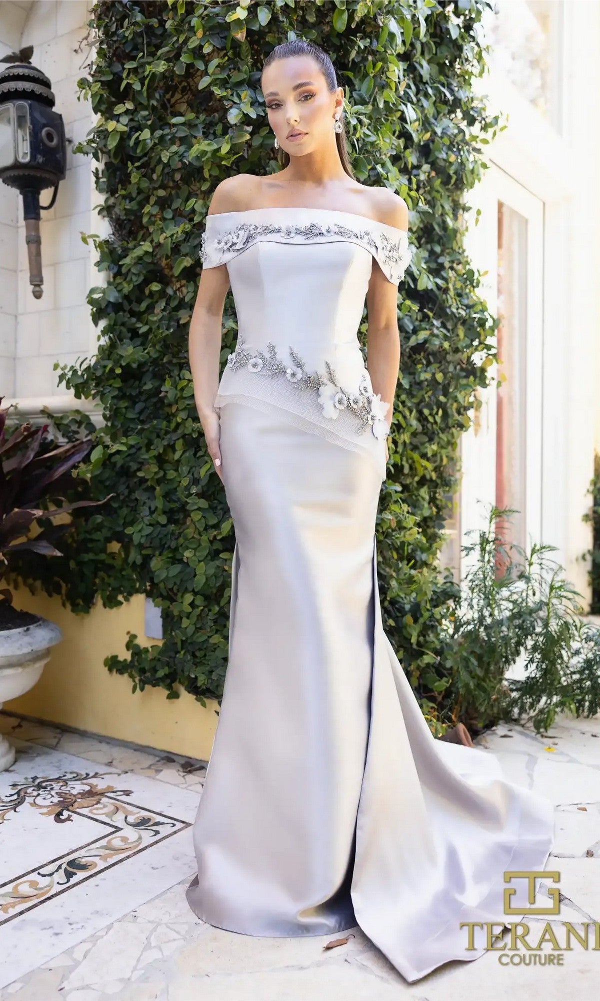 A woman poses outdoors in the Terani 242M3356 Long Formal Dress, a Silver Taupe off-the-shoulder gown with floral and jeweled embellishments, standing gracefully before lush green foliage.
